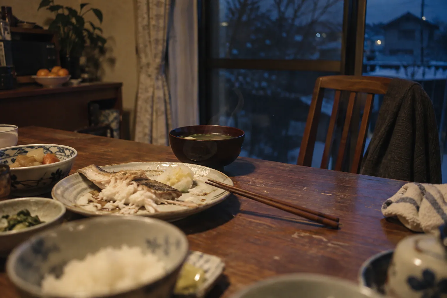 冬の夕食後の食卓。食べかけの白身魚と湯気の残る椀、引かれた椅子