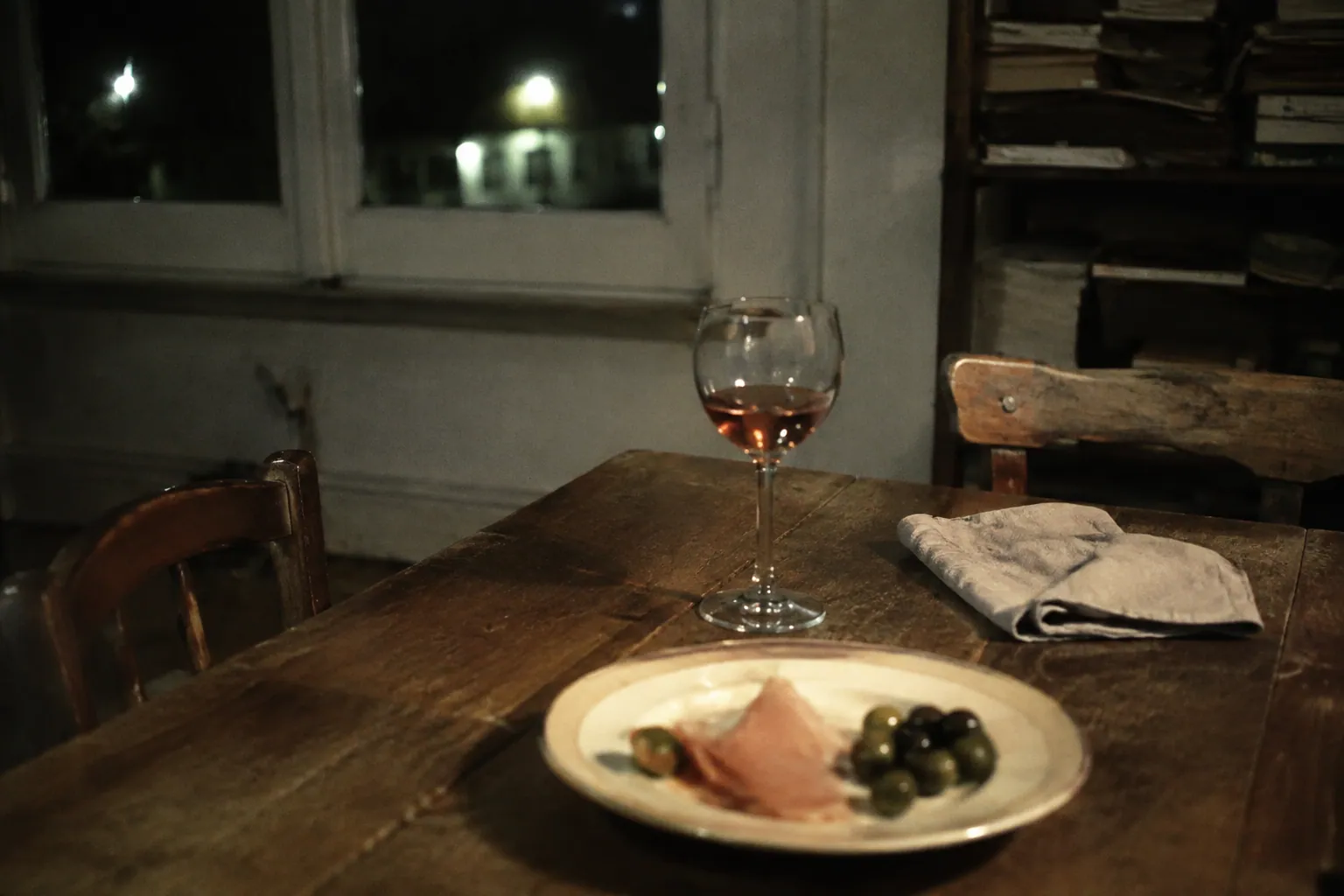 夜の木の食卓に置かれたサーモンピンクのロゼ、生ハムとオリーブ