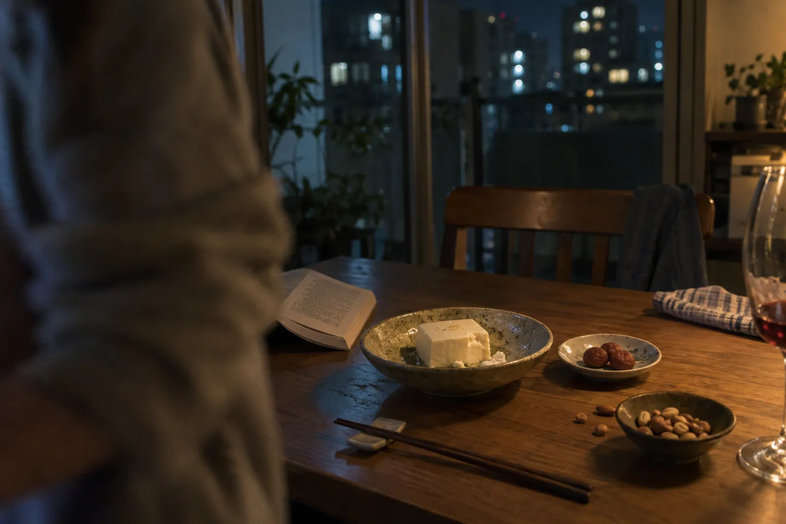 深夜の食卓に、冷奴、梅、ナッツ、伏せた本と飲みかけのグラスがある
