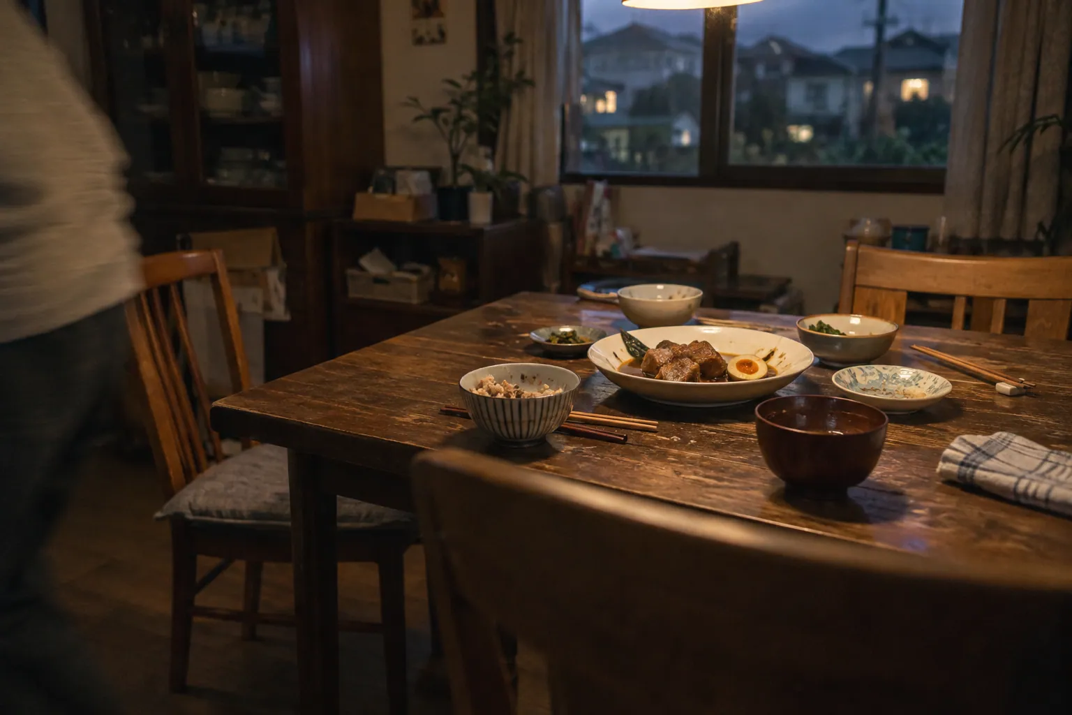 角煮と混ぜご飯が少し残る夕食後の食卓と、引かれた椅子、画面の端を離れる人の肩