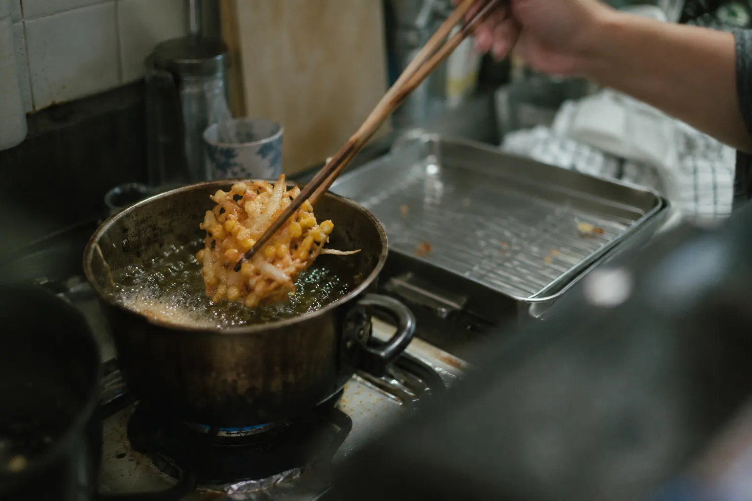 使い込まれた揚げ鍋から、とうもろこしのかき揚げを菜箸で上げる途中の手元