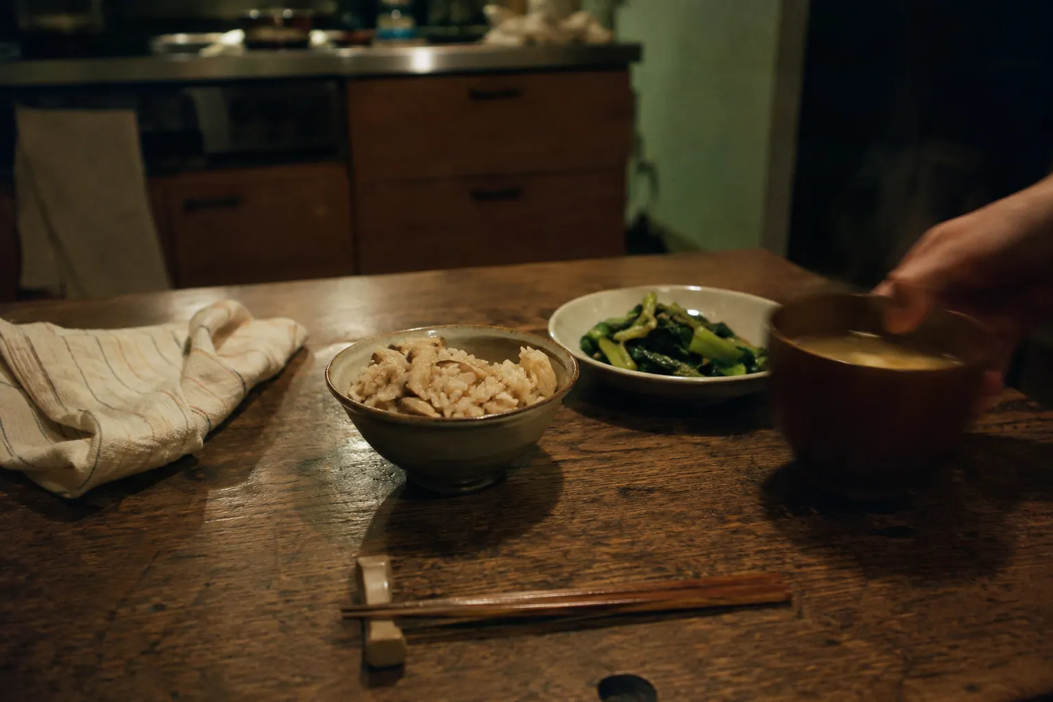 夜の木の食卓に並ぶきのこご飯、青菜、味噌汁と箸