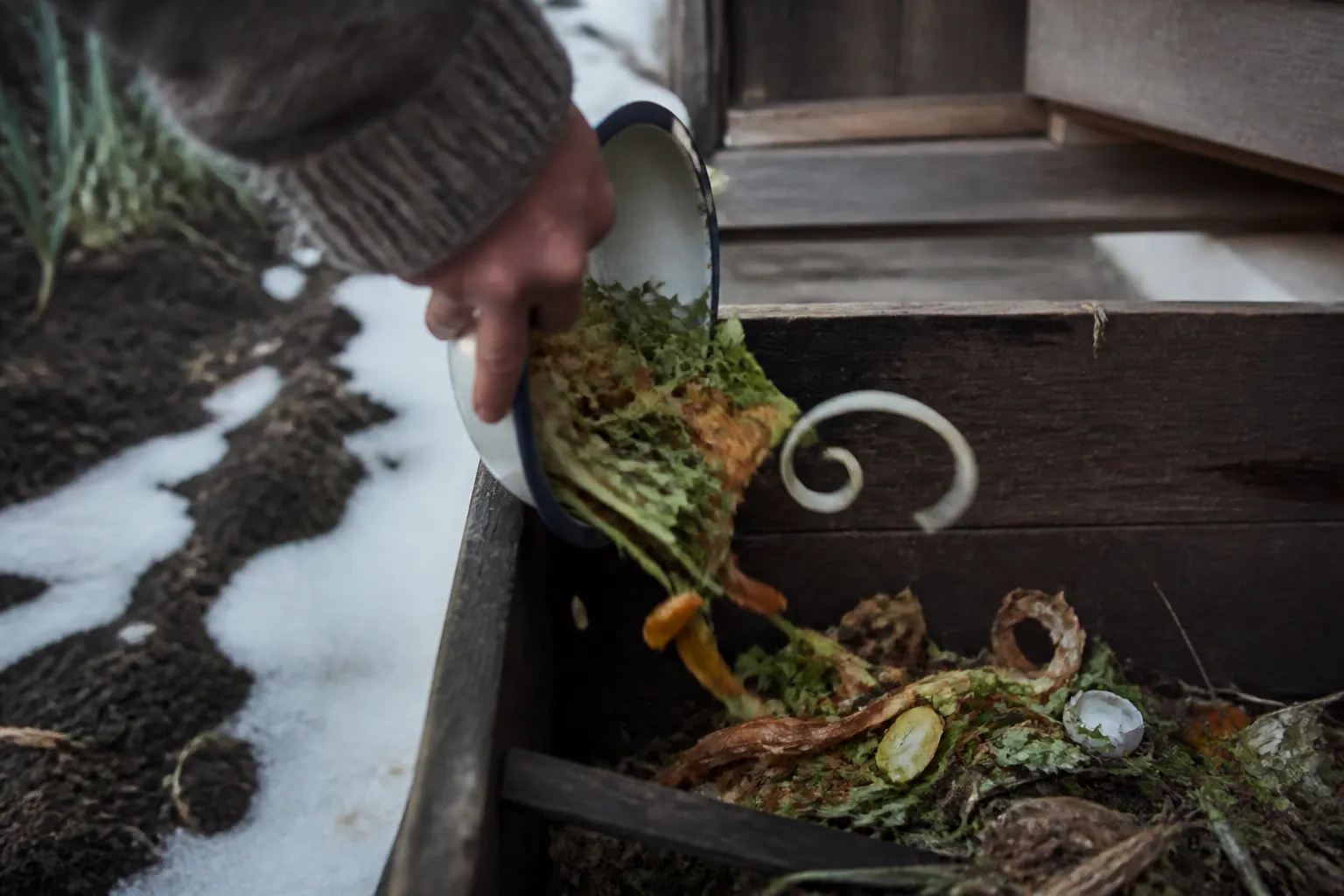 雪の残る家の裏で野菜くずを木の堆肥箱へ戻す手元