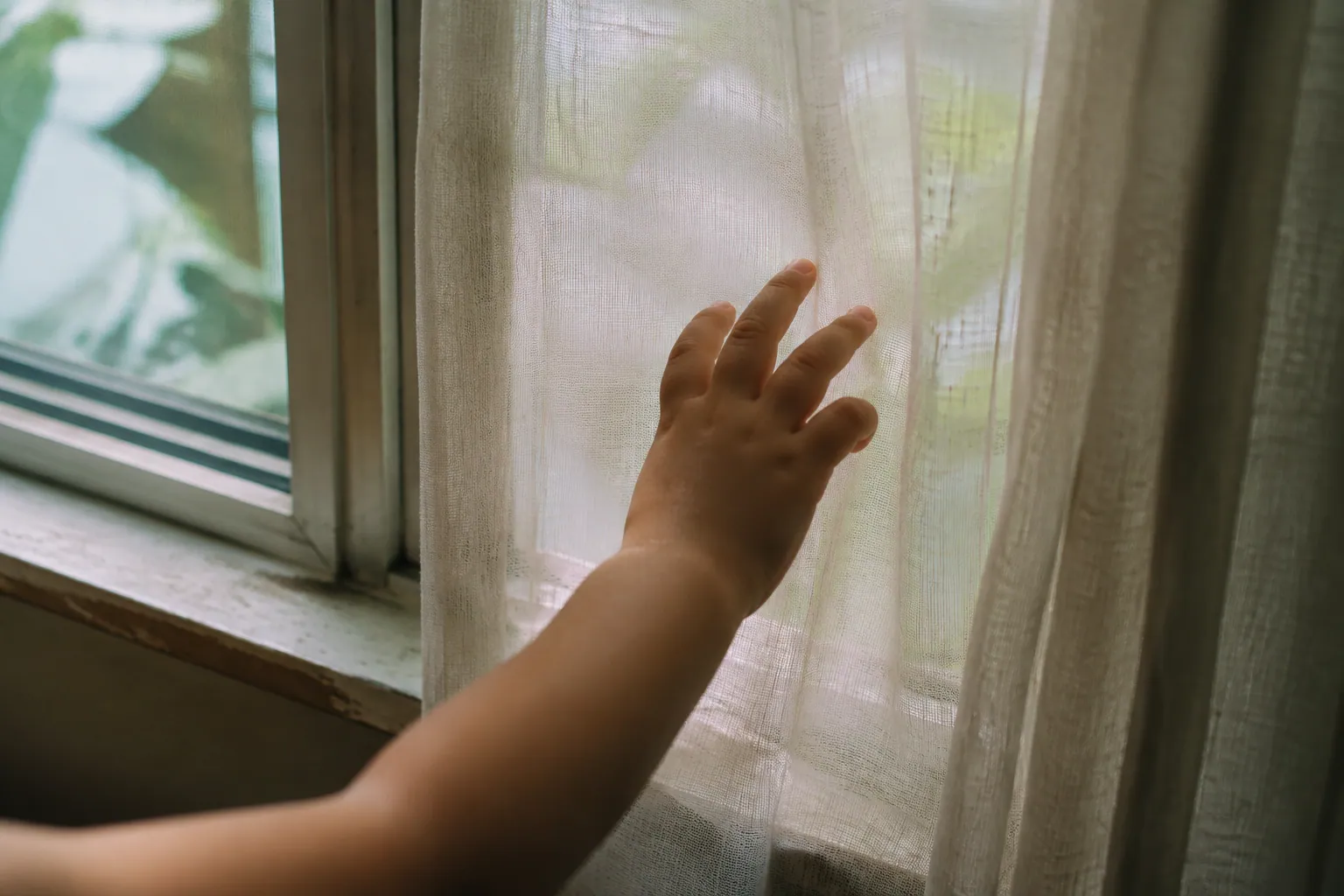 薄いカーテン越しの朝の光へ伸ばされた子どもの手
